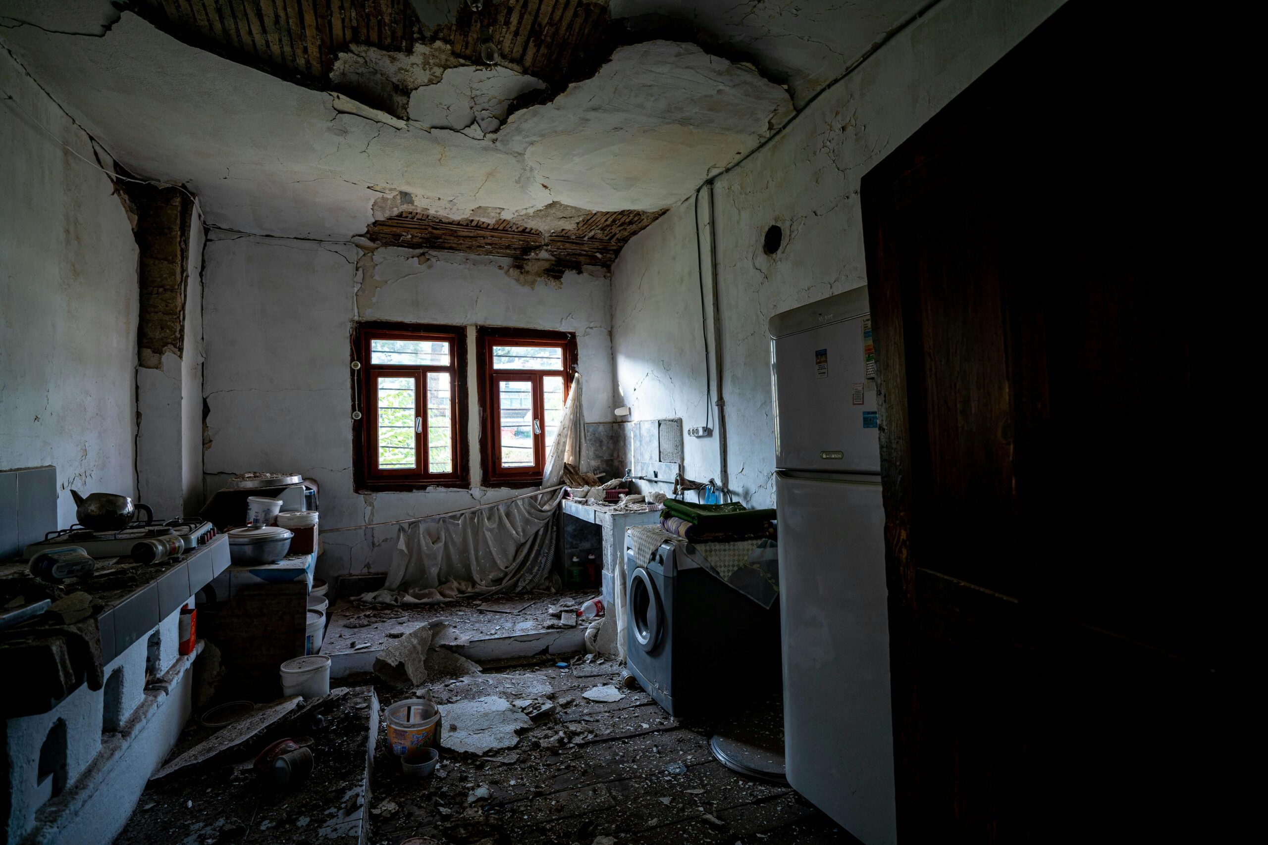 A dilapidated kitchen interior with a collapsing ceiling and scattered debris.